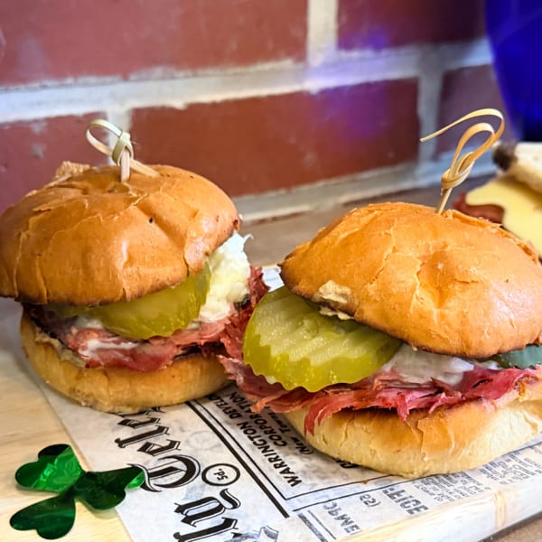 Two mini sandwiches with pastrami, pickles, and sauce on a newspaper-patterned paper with a shamrock.