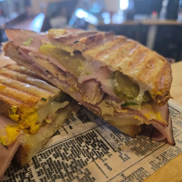 A Cuban sandwich, cut in half, with melted cheese, ham, pickles, and mustard on newspaper.