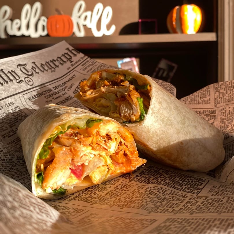 Two Uplifted Bistro wraps, filled with chicken and lettuce, on newspaper with "hello fall" decor.