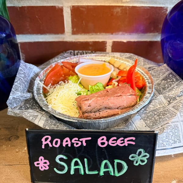 A roast beef salad with shredded cheese, red peppers, and dressing in a foil container.