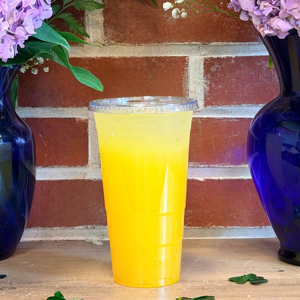 A vibrant yellow drink in a clear plastic cup, flanked by blue vases with purple flowers, against a brick wall.