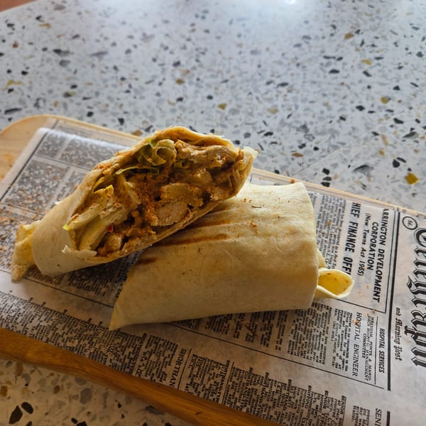 Two chicken shawarma wraps, one cut in half, rest on newspaper on a wooden board.