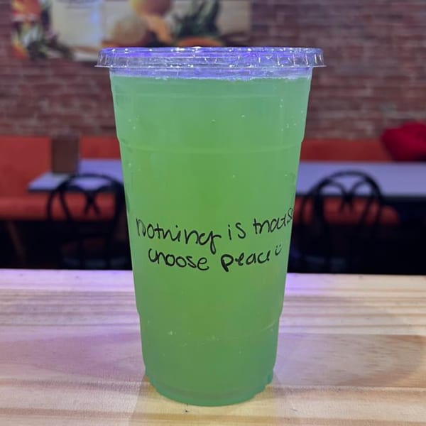 A vibrant green drink in a clear plastic cup with "Nothing is more precious, choose peace" written on it.