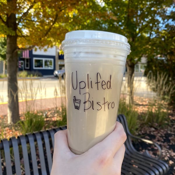 Hand holding an Uplifted Bistro bubble tea cup outdoors with autumn trees and buildings in the background.