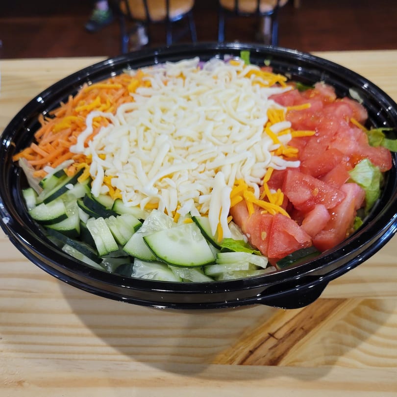 A vibrant Uplifted Bistro salad with shredded cheese, carrots, cucumbers, and tomatoes in a black bowl.