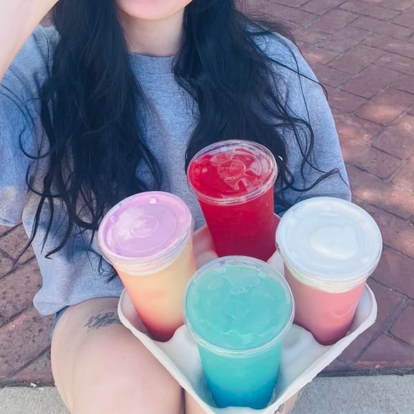A person holds a carrier with four colorful iced drinks: red, orange, blue, and pink.