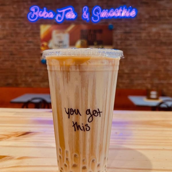 Boba tea with "you got this" written on the cup, against a brick wall with a neon sign.