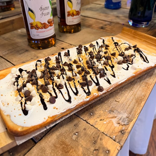 A long flatbread dessert topped with white frosting, chocolate chips, and chocolate drizzle on a wooden board.