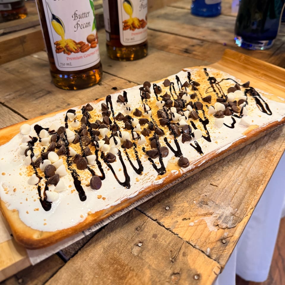 A long flatbread dessert topped with white frosting, chocolate chips, and chocolate drizzle on a wooden board.