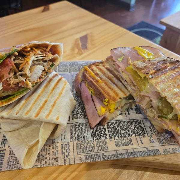 A grilled chicken wrap and a Cuban sandwich, both cut in half, on a newspaper-lined wooden board.
