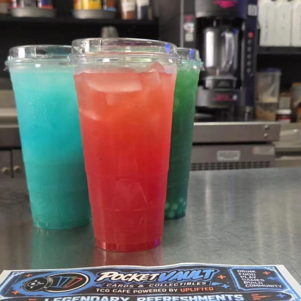 Three colorful iced beverages in red, blue, and green cups sit on a café counter beside a Pocket Vault promotional sign.