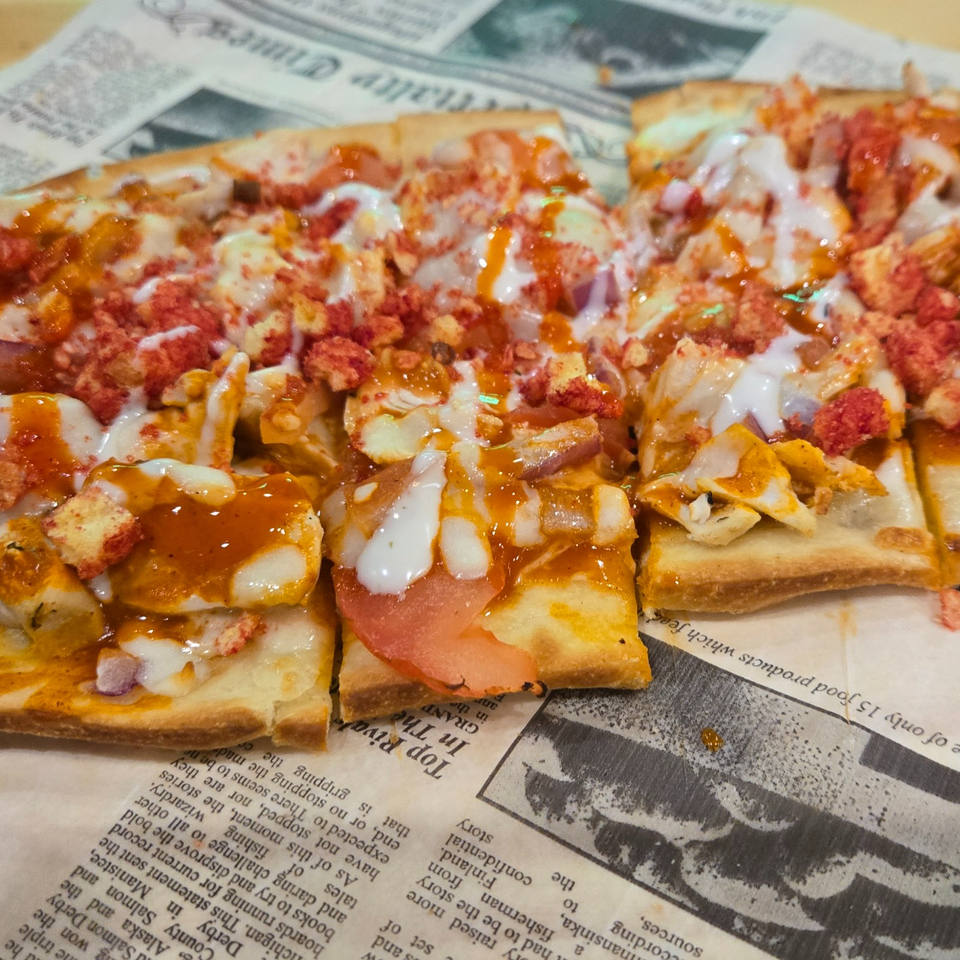 Slices of cheesy pizza topped with vegetables, crispy red crumbs, and drizzled orange and white sauces on newspaper.