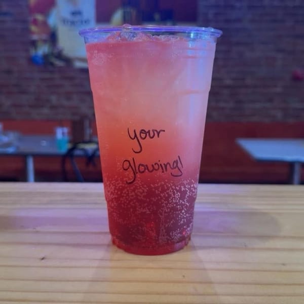 A pink Uplifted Bistro drink with "your glowing!" written on it, on a wooden table.