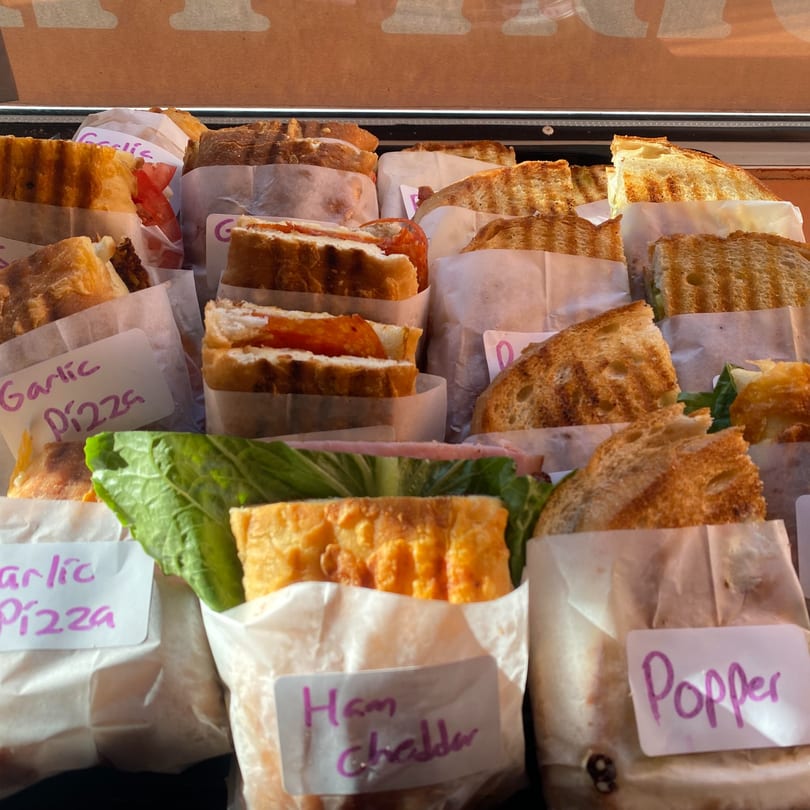 Assortment of Uplifted Bistro cafe sandwiches, including "Ham Cheddar" and "Garlic Pizza," wrapped in paper.